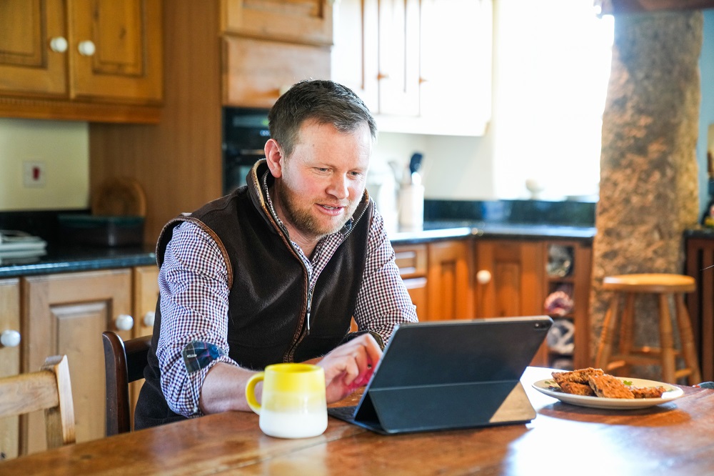 Person using a tablet at a wooden kitchen table with a mug and a plate of biscuits nearby.