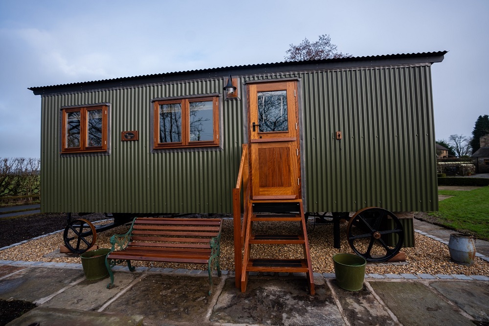 Green corrugated shepherd’s hut on wheels with wooden steps and windows.