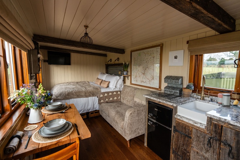 Interior of a shepherd's hut with a bed, sofa, kitchenette, and dining table by the window.